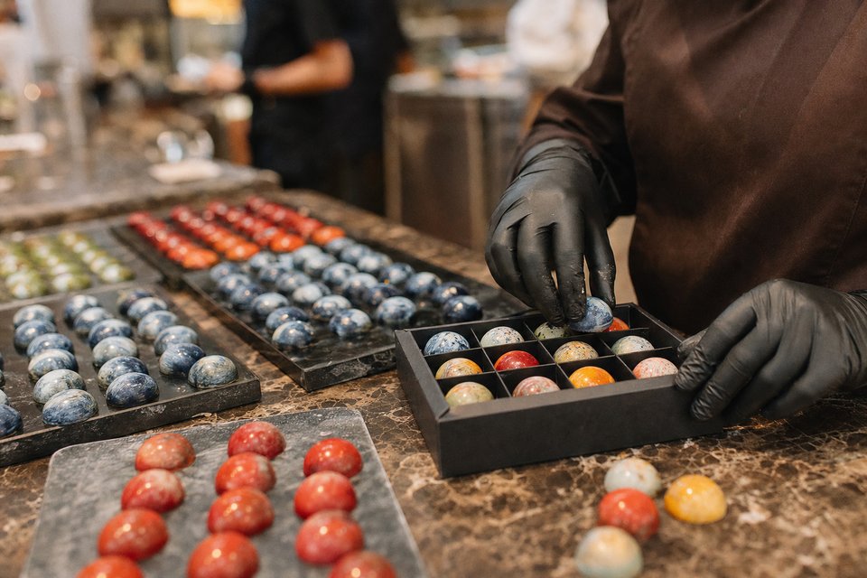 Handen in handschoenen leggen beschilderde bonbons met gemarmerde kleuren in een doosje, met trays bonbons op de werkbank (met AI gemaakte afbeelding)