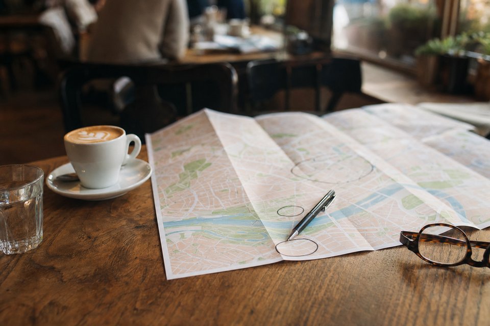 Opgevouwen stadsplattegrond op een cafétafel naast een flat white en een leesbril, met een pen die cirkels zet (met AI gemaakte afbeelding)