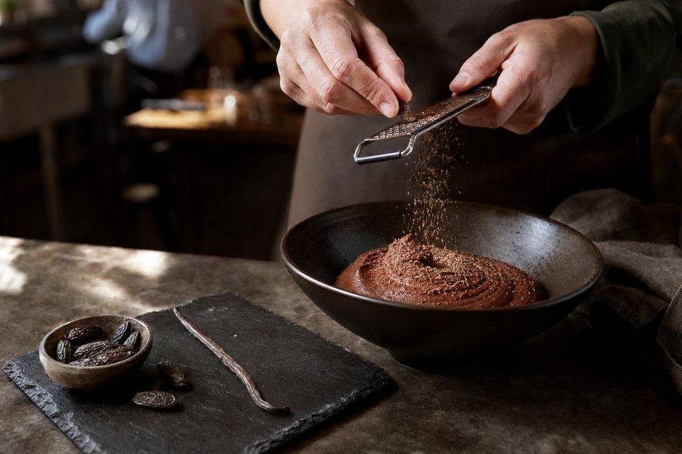 Een tonkaboon wordt geraspt boven een kom donkere chocolademousse, met hele bonen en een vanillestokje op een leisteen (met AI gemaakte afbeelding)