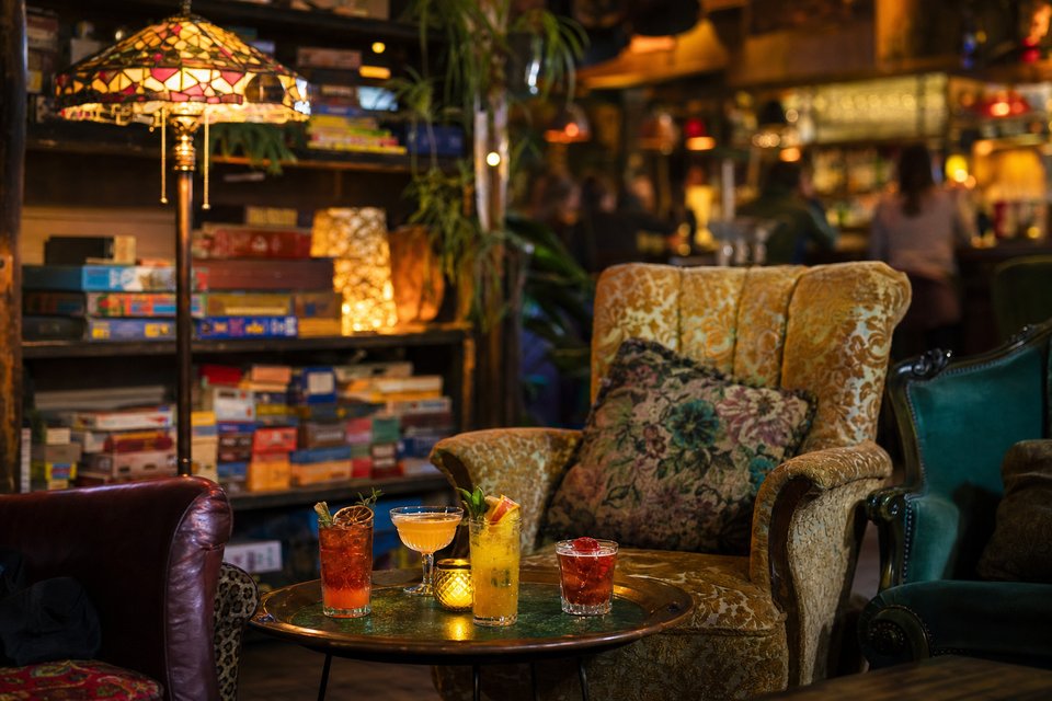 Knus bar-interieur met verschillende vintage fauteuils, tiffanylampen, bordspellen op planken en cocktails op een lage tafel (met AI gemaakte afbeelding)