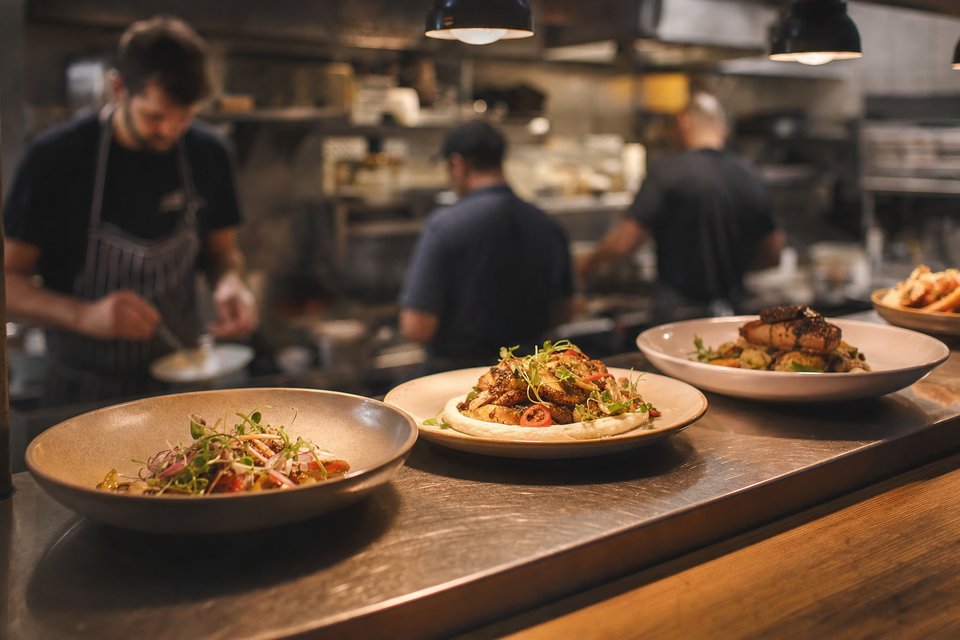 Drukke restaurantkeuken gezien door de pass, koks aan het fornuis en een rij opgemaakte borden op de toonbank (met AI gemaakte afbeelding)